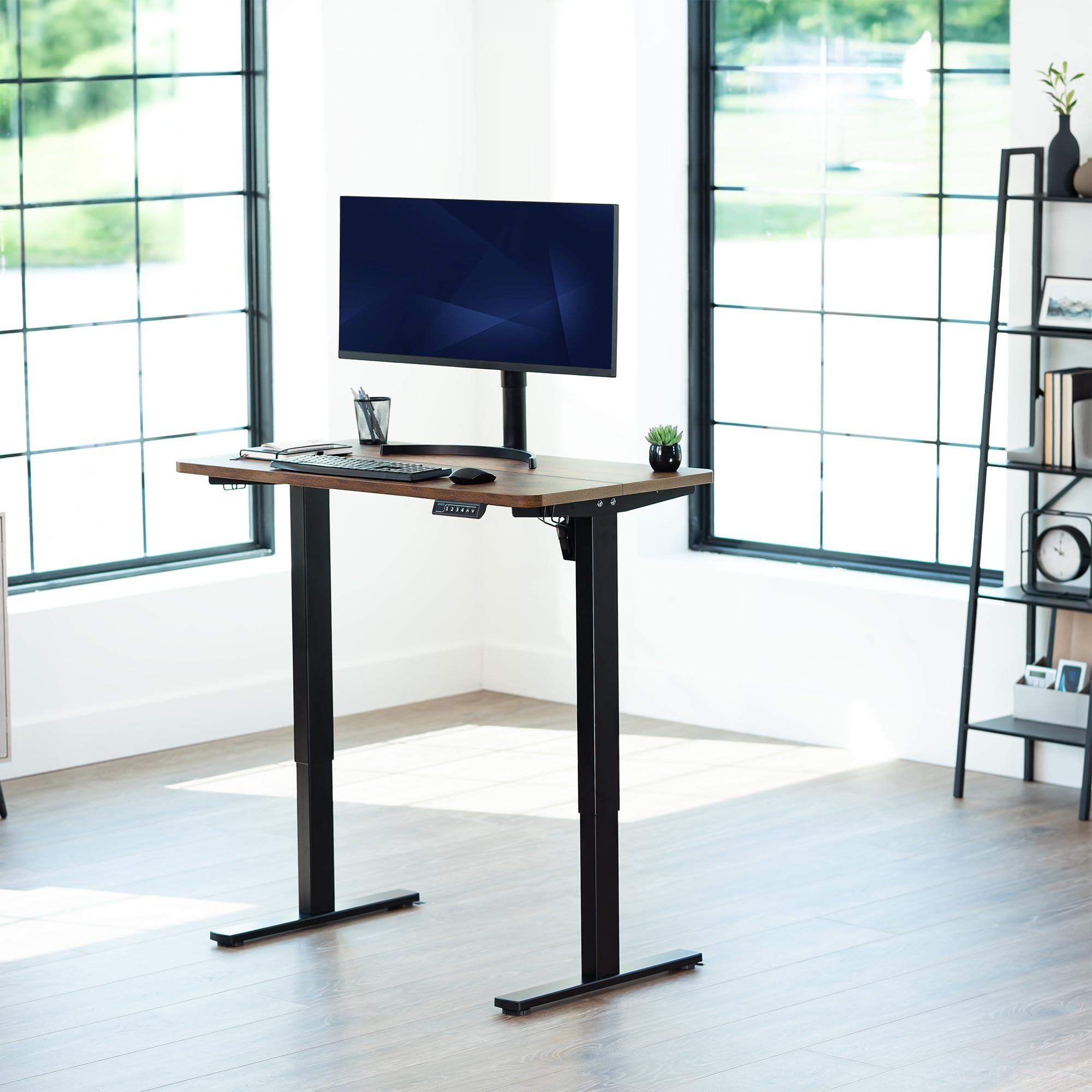 Rustic, adjustable height desk with a computer setup in a bright room with large windows.