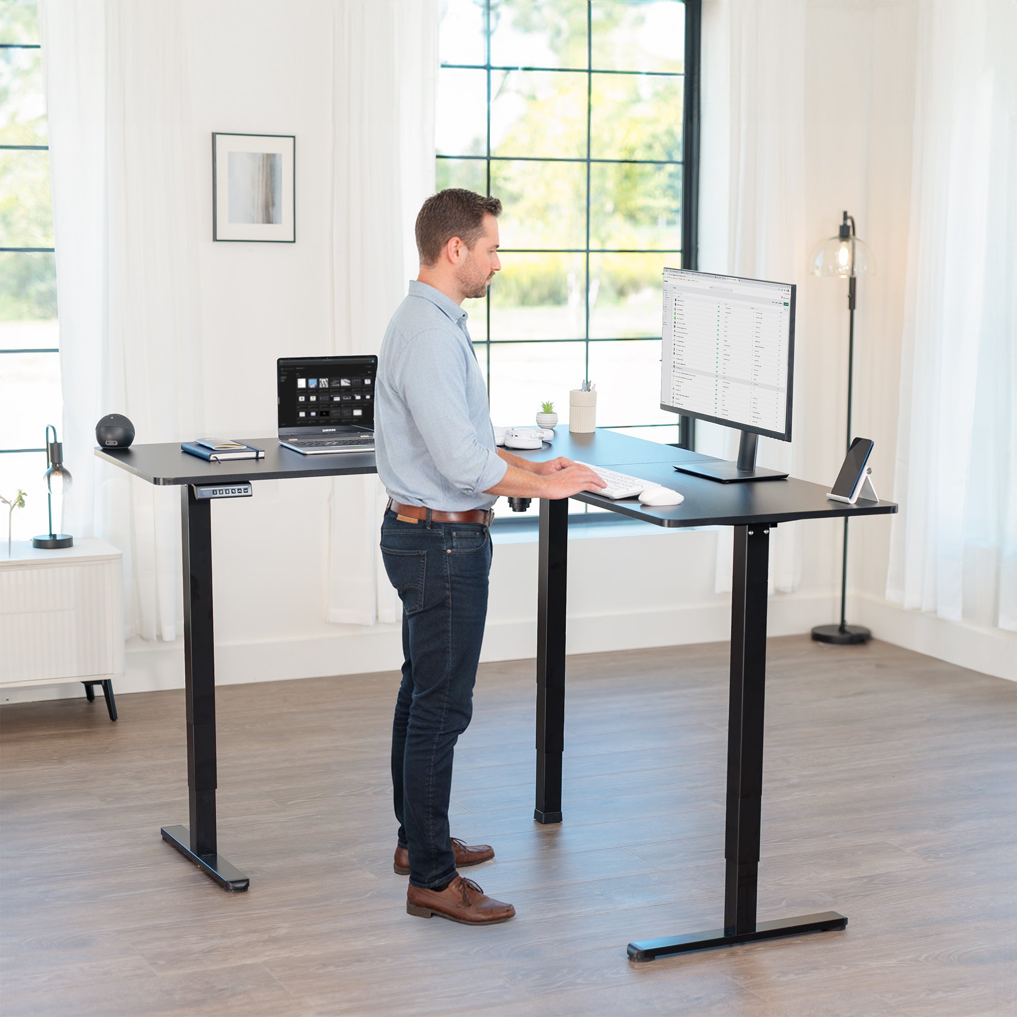 Man using a standing l-shaped desk in a bright room with large windows.