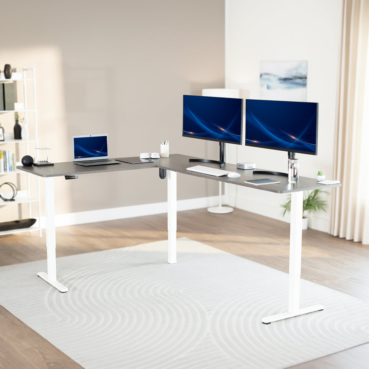 Modern office setup with a standing l-shaped desk, laptop, and two monitors in a bright room.