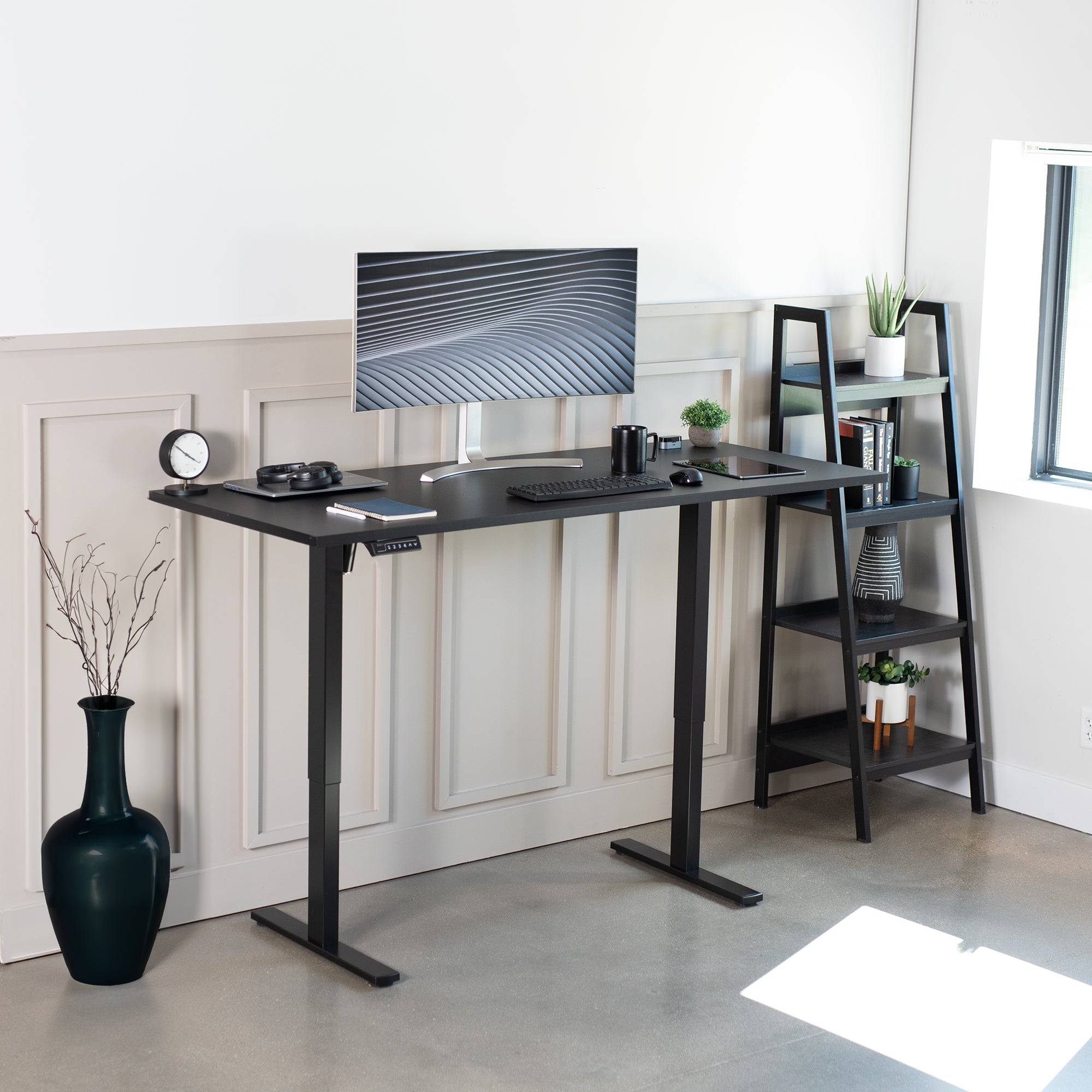 Black adjustable desk in a modern office setting with a monitor and shelves.