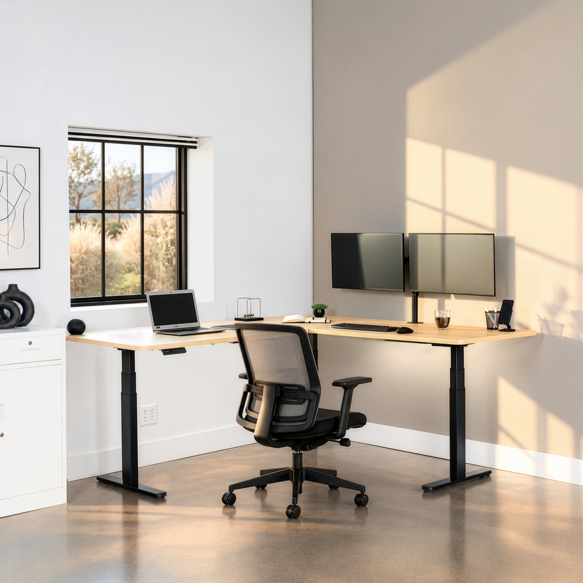 A modern L-shaped standing desk in a bright home office setup, featuring a black mesh office chair, dual computer monitors, a laptop, and a window with natural sunlight casting shadows on the wall.