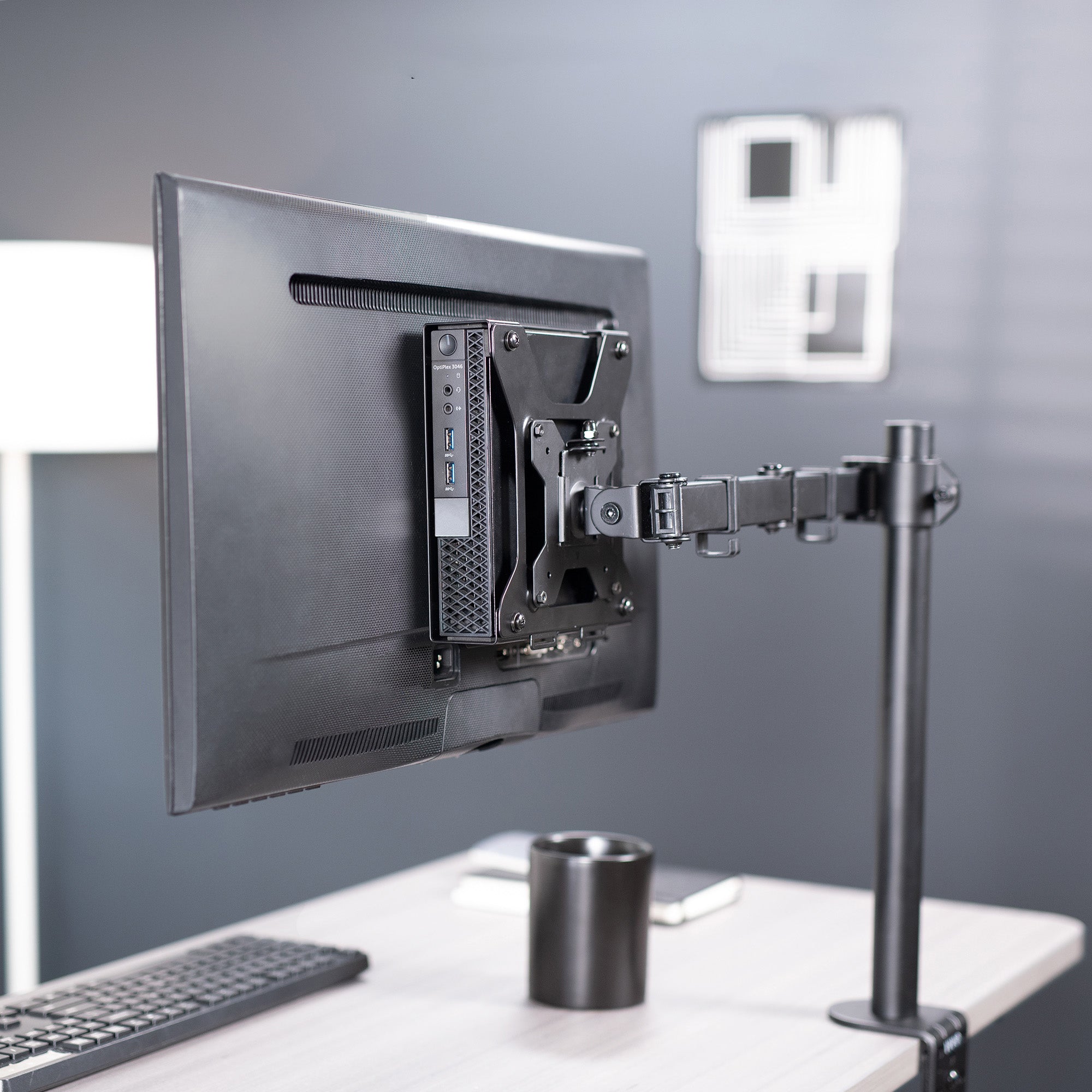 Monitor mounted on a flexible arm stand with a Mount Designed for Dell OptiPlex Micro on the back and a keyboard and cup on a desk.