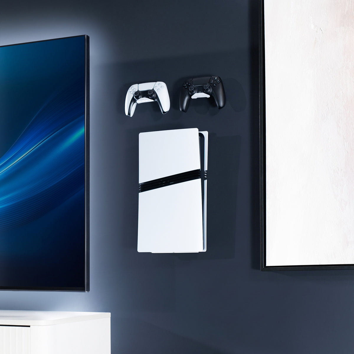 A white PlayStation 5 console with a black accent strip is mounted on a dark wall, flanked by two game controllers, with a TV and framed wall art visible in a modern room setting.