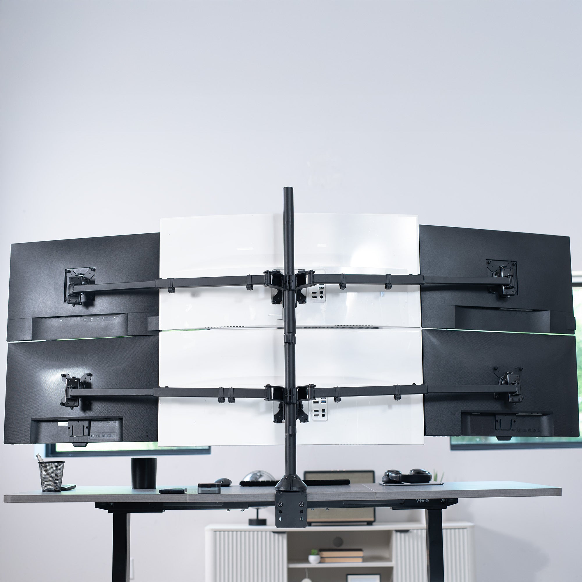 Multiple computer monitors on a desk with a monitor stand against a white wall.
