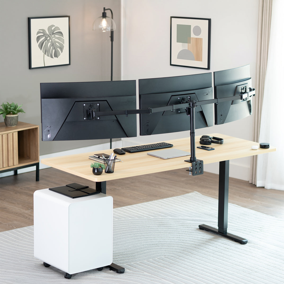 A modern standing desk setup in a bright home office, featuring a light wood desktop, black T-style legs, dual curved monitor arms holding three monitors, a white pedestal storage unit, and desk accessories on a light rug.