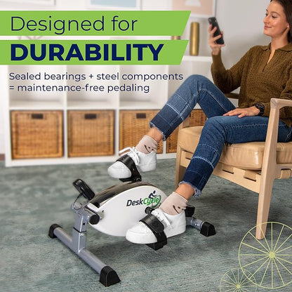 A woman sits on a wooden chair in a home office setting, using a white and black desk exercise bike. The image features promotional text overlays highlighting the bike's durability and features.