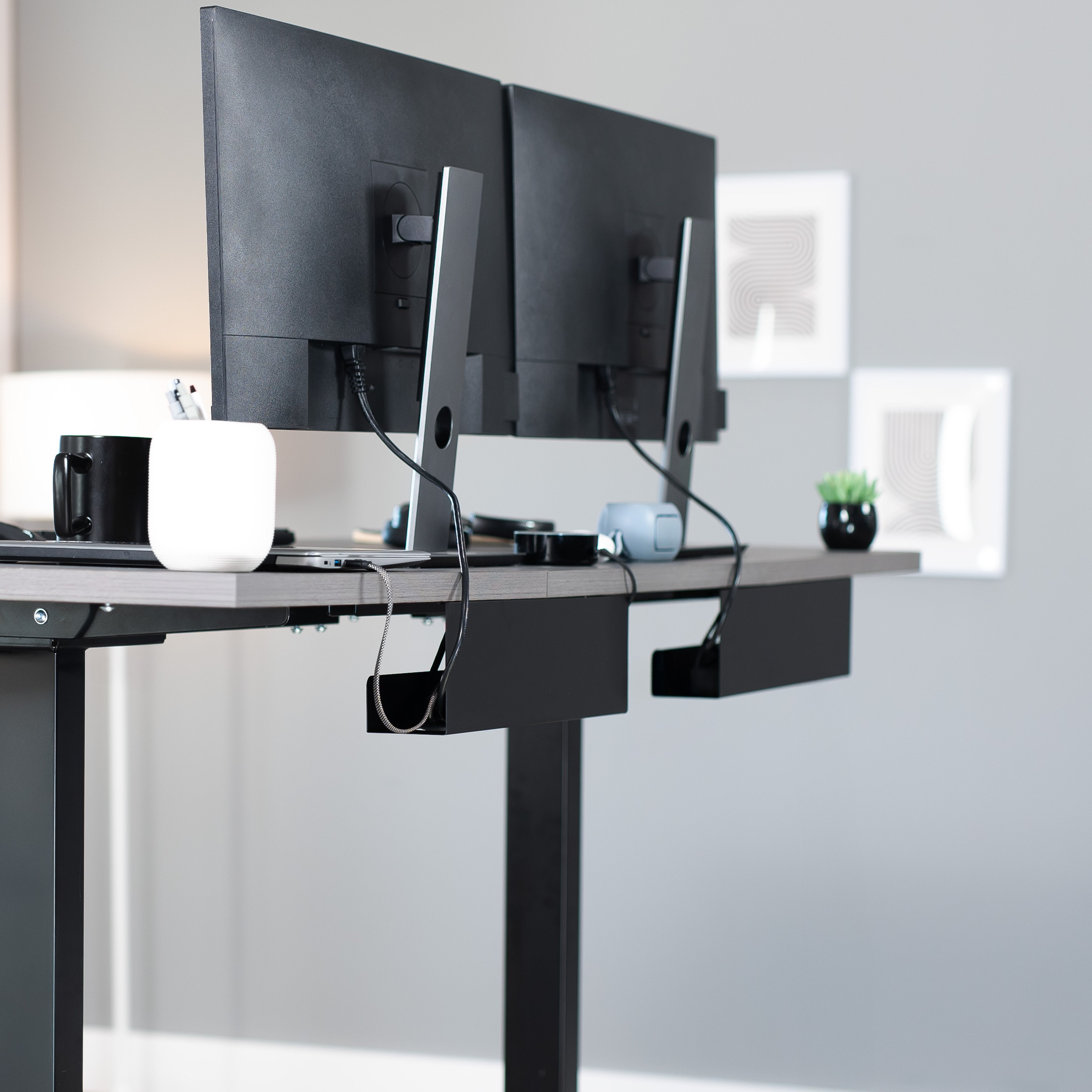 Modern office setup with a black desk and dual monitors on a light gray wall background with cable management trays below the desk.