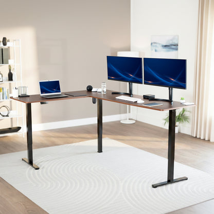 Modern office setup with a standing l-shaped desk, laptop, and two monitors in a bright room.