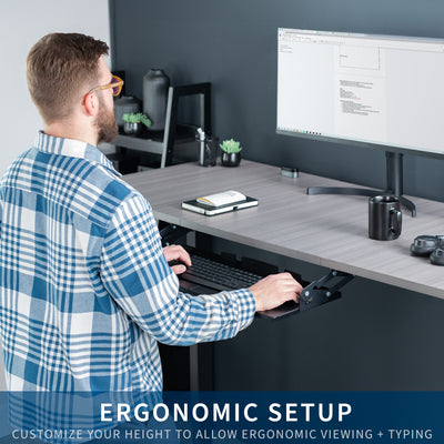 Man working at an ergonomic sit-to-stand desk with an under-desk mounted keyboard tray.