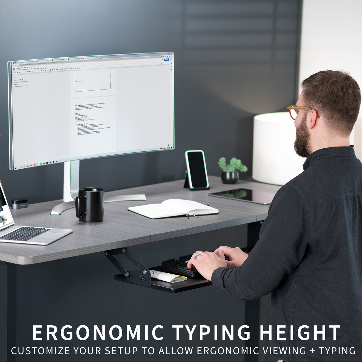 Man working at an ergonomic sit-to-stand desk with an under-desk mounted keyboard tray.