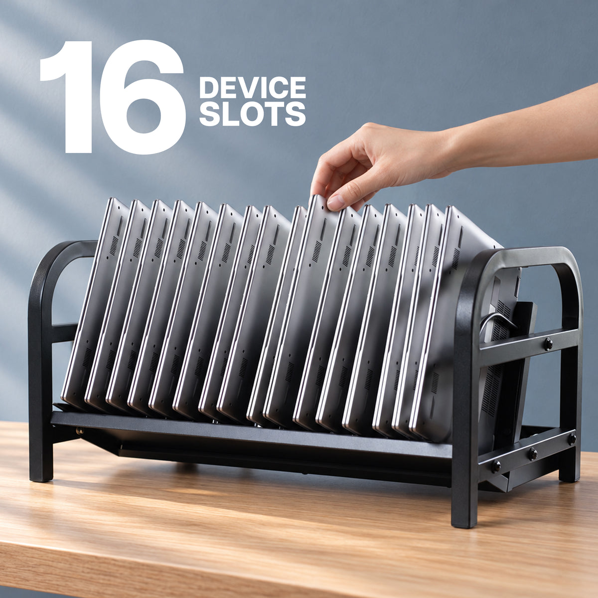 A black metal 16-device laptop stand holding multiple slim silver laptops, with a hand inserting one device. The image includes the text '16 DEVICE SLOTS' and is set on a wooden desk against a blue-gray background.