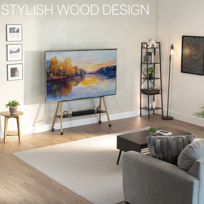 A modern living room scene showing a large wall-mounted TV on a slim wooden rolling stand with casters. The room has light wood flooring, a cream rug, a gray sofa, a black coffee table, a floor lamp, and framed wall art.