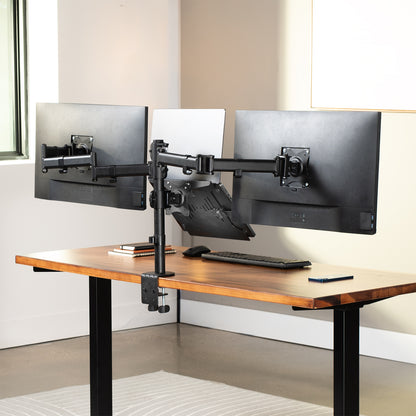 A dual monitor arm with laptop stand holding two black computer monitors and a laptop above a wooden desk in a bright home office setting.