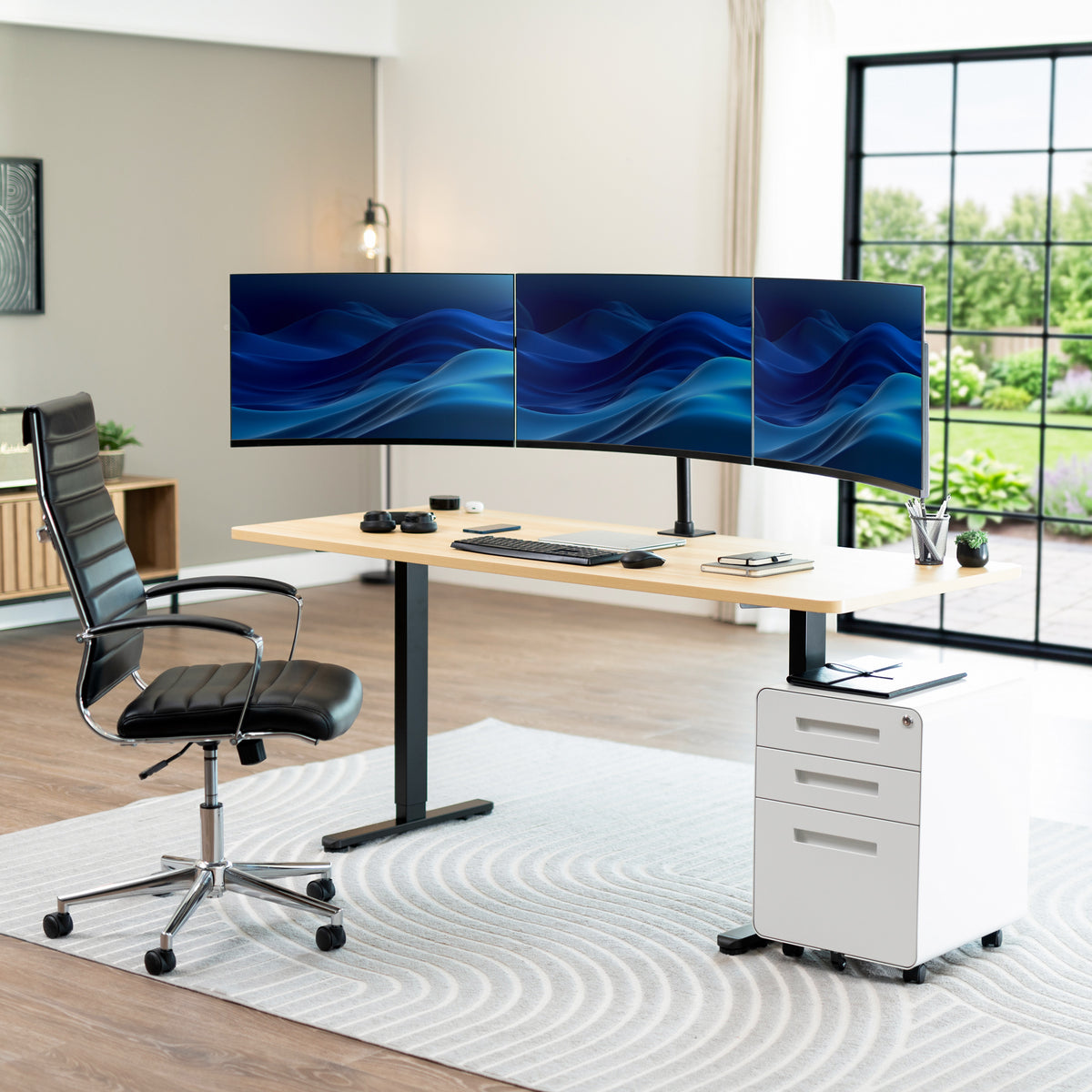 A modern home office setup featuring a light wood standing desk with a black metal frame, a curved triple-monitor desktop setup, a black leather office chair, a white rolling filing cabinet, and a gray patterned rug in a bright room with a large window.