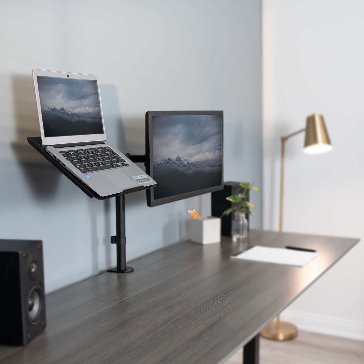 A laptop stands on the same pole as a single monitor mount holding the laptop screen and monitor screen at similar levels.
